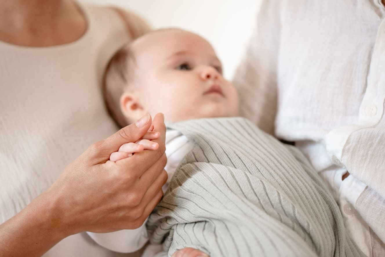 Eine Nahaufnahme eines Babys, das in den Armen eines Erwachsenen ruht, während ein anderer Erwachsener sanft die Hand des Babys hält. Die Erwachsenen tragen helle Kleidung, was eine sanfte und beruhigende Atmosphäre schafft.